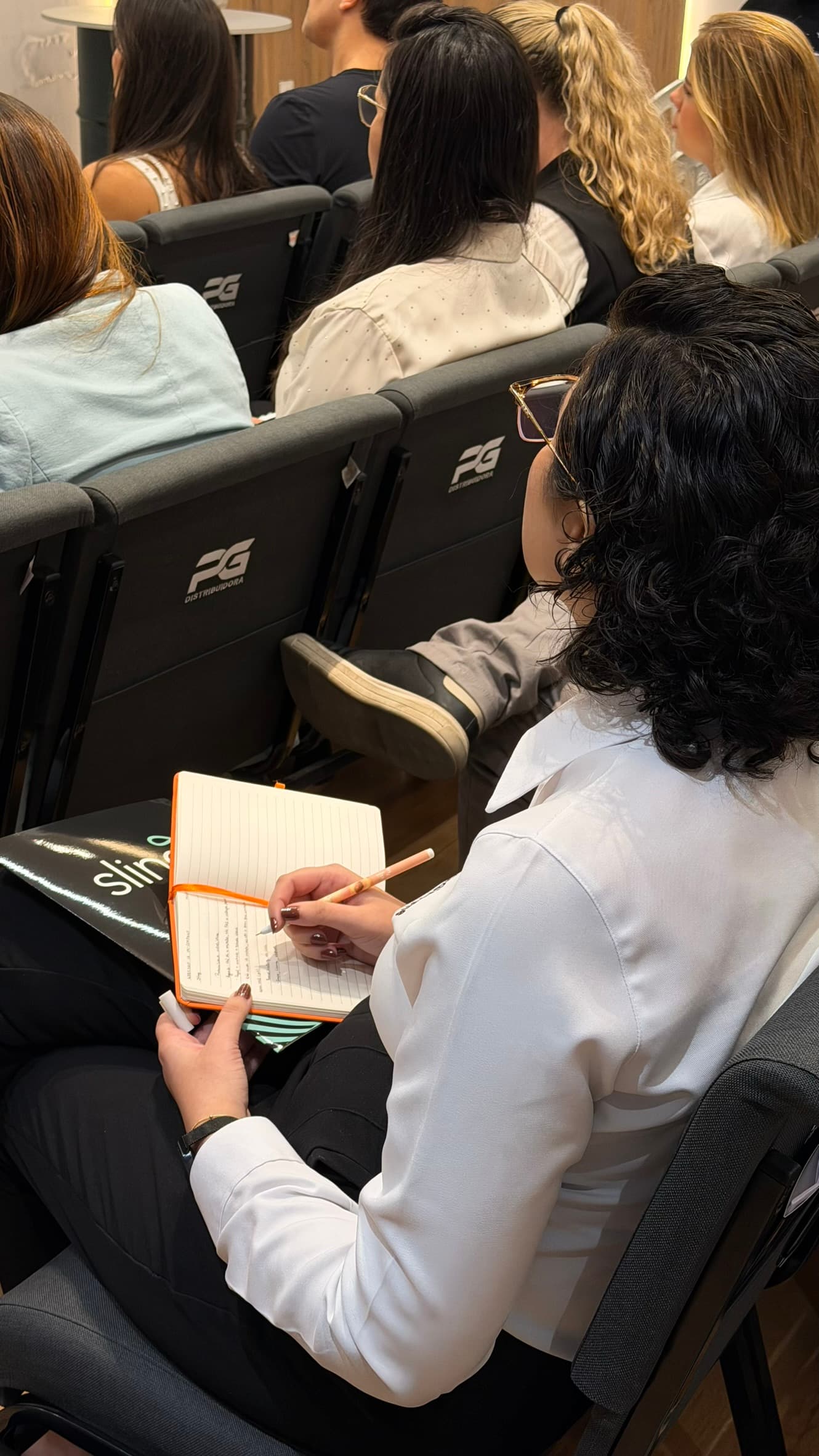 Participante anotando em um caderno apoiado no colo, no meio da plateia do workshop