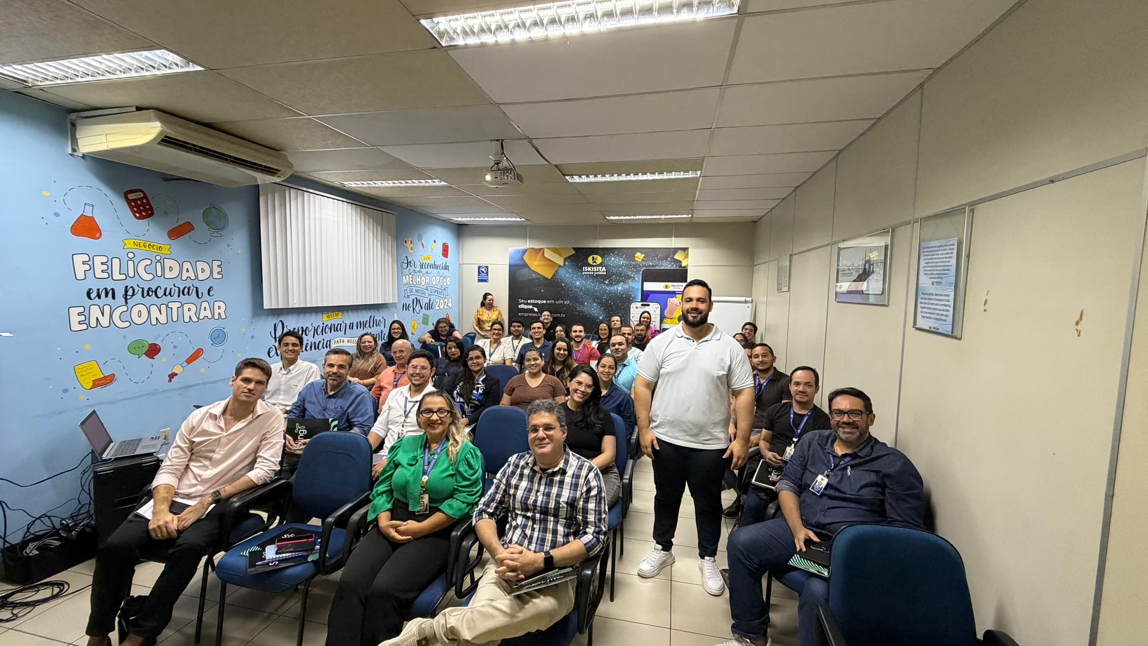 Turma completa reunida na sala de uma empresa, sentada em fileiras, com o instrutor em pé ao centro
