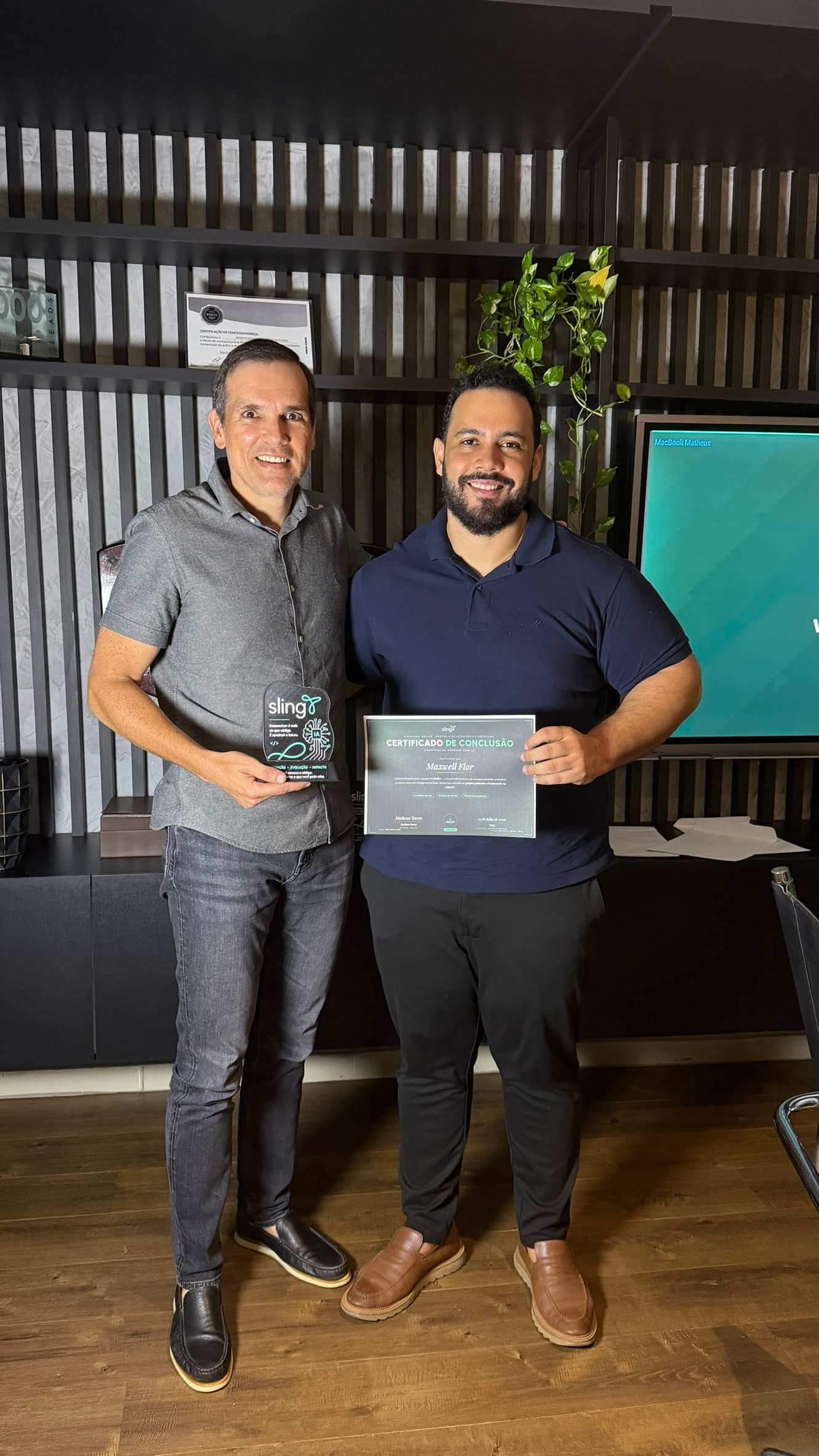 Dupla em pé diante do painel ripado escuro, um segurando o certificado de conclusão e o outro o troféu de acrílico com o logotipo da marca