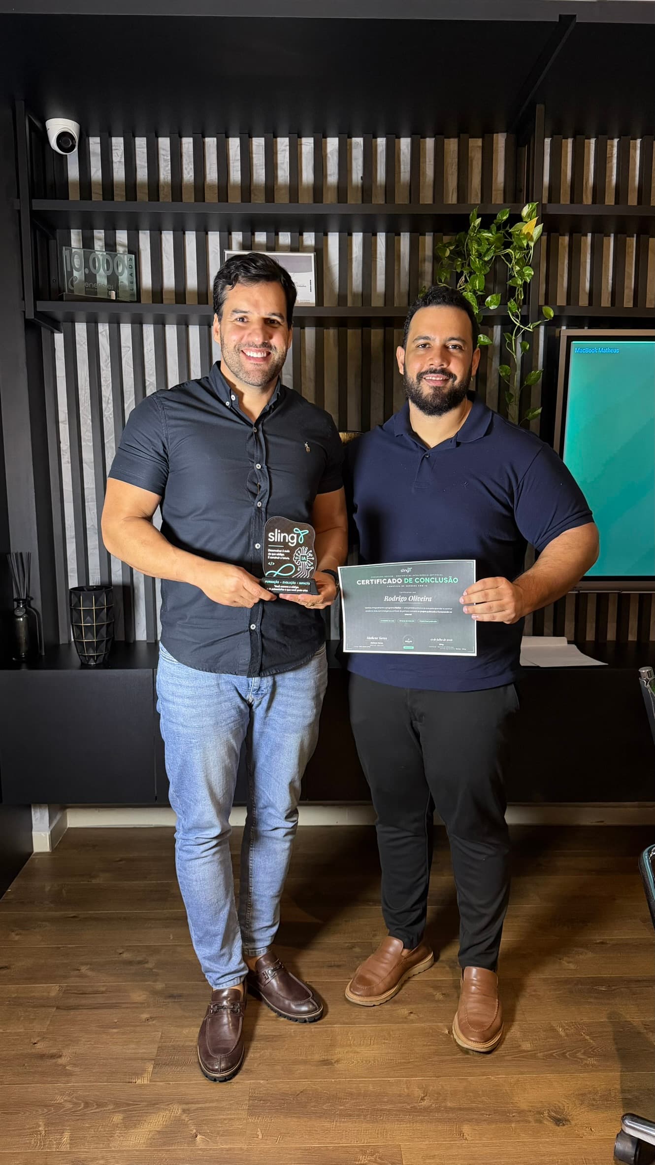 Aluno de camisa escura com o troféu de acrílico ao lado de quem lhe entrega o certificado de conclusão, diante da estante da sala