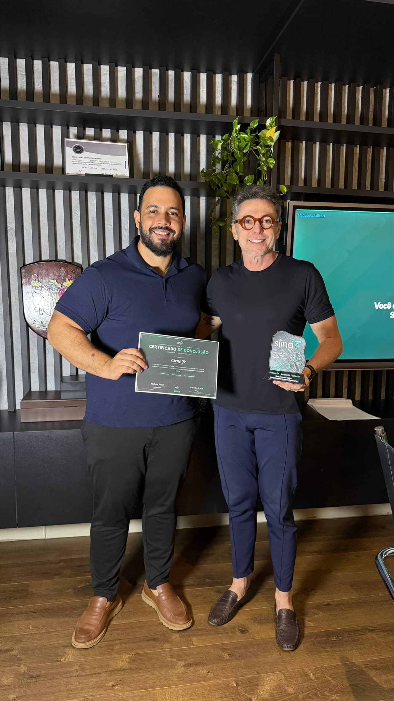 Dois participantes sorrindo diante do telão, um com o certificado de conclusão nas mãos e o outro com o troféu de acrílico