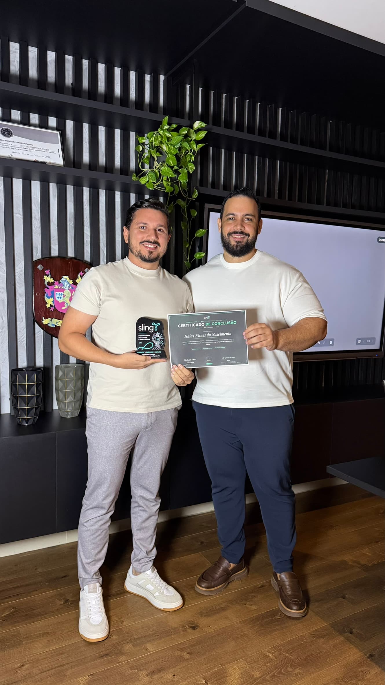Dupla de camiseta clara exibindo o certificado de conclusão e o troféu de acrílico em frente ao telão da sala