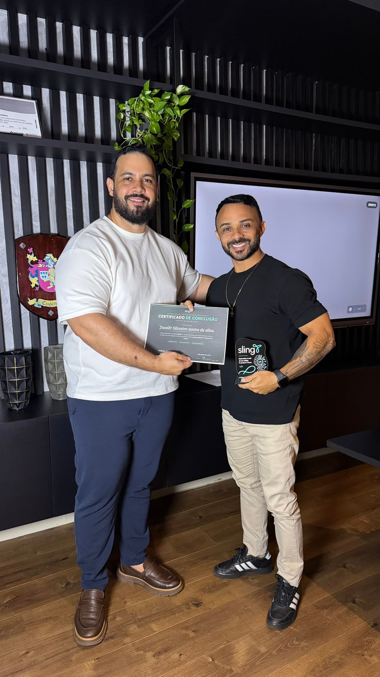 Dois participantes juntos, um com a mão no ombro do outro, exibindo o certificado de conclusão e o troféu de acrílico