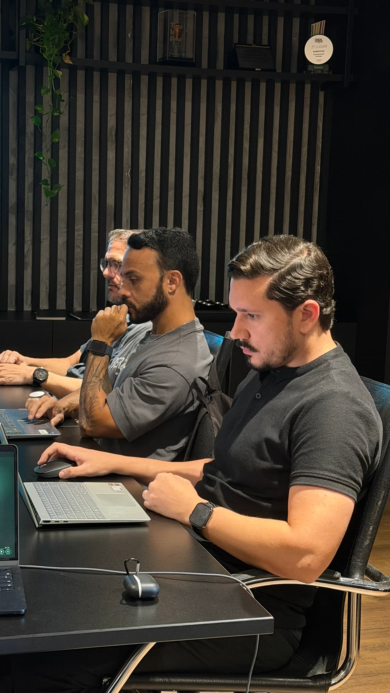 Três participantes enfileirados na mesa de reunião, cada um com a mão no teclado do próprio notebook